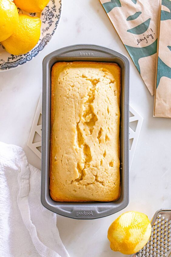 starbucks copycat lemon loaf, A loaf of lemon bread in a baking pan on a white surface surrounded by lemons and a kitchen towel