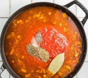 stracotto italian pot roast, Black pot of soup with diced vegetables tomatoes herbs and a bay leaf on a light tiled surface