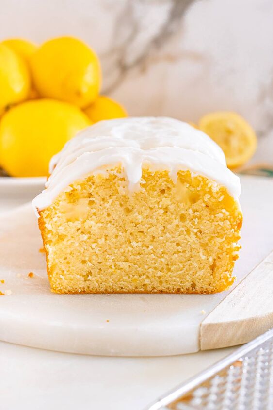 starbucks copycat lemon loaf, A sliced lemon loaf cake with white icing shown on a marble board with fresh lemons in the background