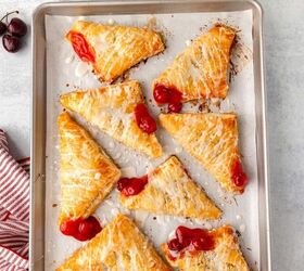 cherry turnovers, A baking tray with eight glazed cherry turnovers arranged in rows Some turnovers have cherry filling oozing out A bowl of fresh cherries and two bottles of milk in a wire holder are nearby on a light colored countertop with a striped cloth