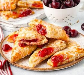 cherry turnovers, A plate of glazed cherry turnovers with flaky pastry and visible cherry filling A white bowl filled with fresh cherries sits nearby and a red striped towel is draped beside the plate on a light surface