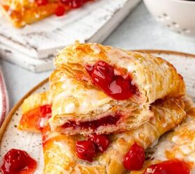 cherry turnovers, Close up of flaky cherry turnovers on a speckled plate drizzled with glaze and filled with bright red cherries A bowl of fresh cherries and more turnovers are visible in the background on a white napkin