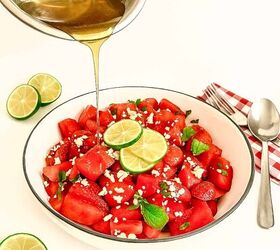 refreshing watermelon strawberry salad recipe, Honey lime dressing being poured onto watermelon strawberry salad with feta and mint in a white bowl styled with lime slices and red gingham napkin