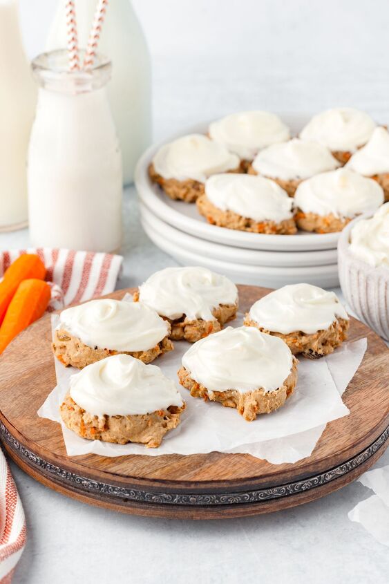 carrot cake cookies, Frosted carrot cake cookies arranged on a wooden platter and a plate in the background A bowl of cookies two bottles of milk a small stack of plates and sliced carrots are also visible
