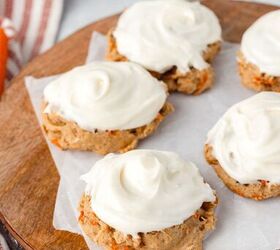 carrot cake cookies, Four carrot cake cookies topped with creamy white frosting are arranged on a wooden board lined with parchment paper A striped cloth and additional cookies can be seen in the background