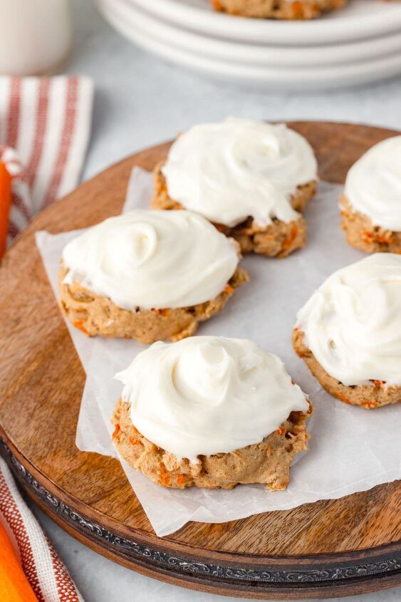carrot cake cookies, Four carrot cake cookies topped with creamy white frosting are arranged on a wooden board lined with parchment paper A striped cloth and additional cookies can be seen in the background