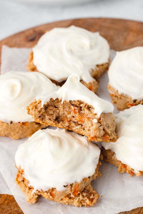 carrot cake cookies, A stack of frosted carrot cake cookies on a wooden surface One cookie is split in half displaying the moist spiced interior with visible pieces of carrot The cookies are topped with smooth cream cheese frosting