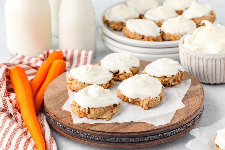 carrot cake cookies, Cookies with white frosting on a wooden board surrounded by two bottles of milk a striped cloth raw carrots and a bowl with more frosting A stack of plates with more cookies is in the background