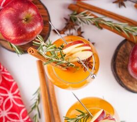 the best fireball apple cider drink for the fall season, Two glasses of Fireball apple cider with apple slices rosemary and cinnamon sticks styled with apples and a red holiday jar in the background