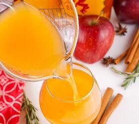the best fireball apple cider drink for the fall season, Apple cider being poured from a glass measuring cup into a round cocktail glass with apples and cinnamon in the background