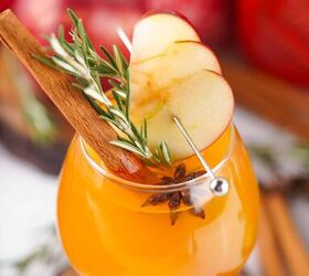 the best fireball apple cider drink for the fall season, Festive Fireball apple cider cocktail in a glass with apple slices rosemary and a cinnamon stick set against a holiday background with apples and spices