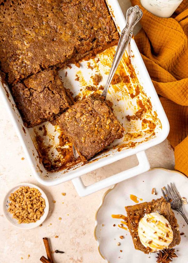 pumpkin dump cake, A white baking dish with pumpkin dump cake one square piece being lifted out with a cake server A plated slice with ice cream and caramel sauce sits nearby with a bowl of chopped nuts and a cinnamon stick on the table