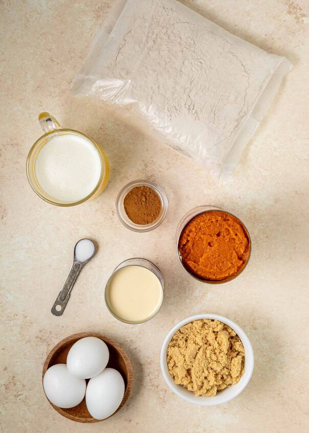 pumpkin dump cake, A flat lay of baking ingredients a bag of cake mix sweetened condensed milk a bowl of brown sugar three eggs a small bowl of melted butter a jar of cinnamon a can of pumpkin puree and a spoon with salt