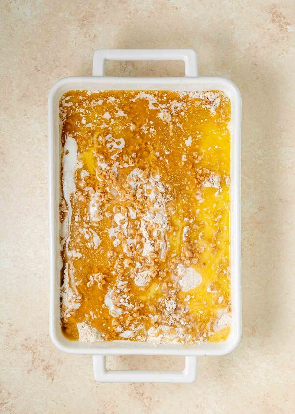 pumpkin dump cake, A rectangular white baking dish filled with an unbaked mixture of yellow melted butter cake mix and some dry ingredients showing a marbled texture on a light colored countertop
