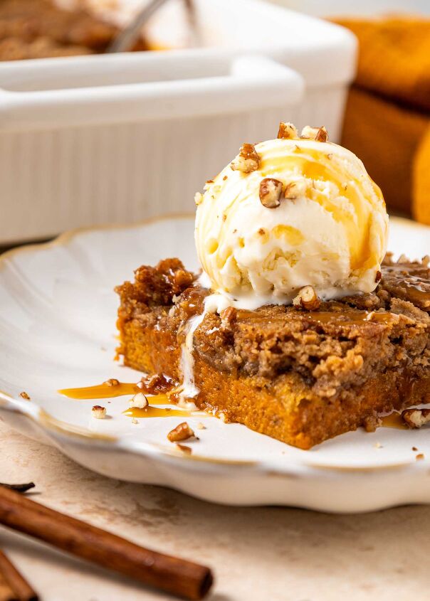 pumpkin dump cake, A slice of pumpkin dump cake topped with a scoop of vanilla ice cream toffee bits and caramel drizzle on a white plate A pan of more dessert and a cinnamon stick are in the background