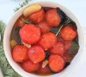 cherry tomato confit with basil and thyme mediterranean style, A bowl from The Sunday Thymes filled with peeled cherry tomatoes fresh basil leaves and garlic cloves marinated in olive oil The bowl rests on a white surface with a green cloth and sprigs of herbs nearby