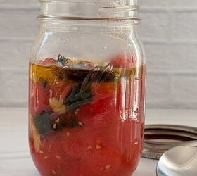 cherry tomato confit with basil and thyme mediterranean style, A glass mason jar filled with cherry tomato confit fresh basil and olive oil sits on a white surface beside its metal lid and a spoon evoking a Mediterranean feel against a light brick background