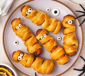 spooky cute air fryer mummy hot dogs kids will love, Crispy mummy hot dog wrapped in crescent dough candy eyes visible plated next to small bowls of dipping sauces