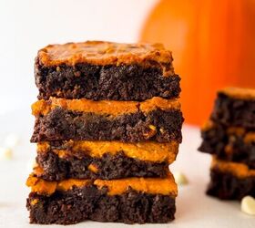 pumpkin cheesecake brownies recipe for fall desserts, Stack of homemade pumpkin cheesecake brownies with rich chocolate layers and creamy pumpkin filling styled with white chocolate chips on parchment and a blurred pumpkin in the background