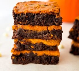 pumpkin cheesecake brownies recipe for fall desserts, Tall stack of pumpkin cheesecake brownies highlighting the swirled chocolate and pumpkin layers on parchment paper with white chocolate chips and a blurred pumpkin behind