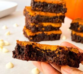 pumpkin cheesecake brownies recipe for fall desserts, Close up of a hand holding a slice of pumpkin cheesecake brownie showing fudgy chocolate and creamy pumpkin layers with more brownies stacked and a pumpkin in the blurred background