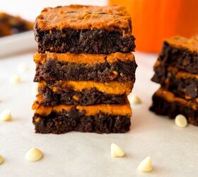 pumpkin cheesecake brownies recipe for fall desserts, Stack of pumpkin cheesecake brownies on parchment showing thick chocolate layers and creamy pumpkin filling with scattered white chocolate chips and a blurred pumpkin in the background