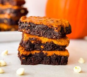 pumpkin cheesecake brownies recipe for fall desserts, Side view of three pumpkin cheesecake brownies stacked on parchment with visible swirled chocolate and pumpkin layers white chocolate chips and a pumpkin in the blurred background