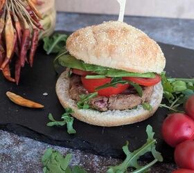 spicy jackfruit and red kidney bean burgers, A plant based bean burger with a jackfruit served in a whole grain gluten free bun The burger is topped with fresh avocado slices a thick slice of tomato and leafy greens likely arugula adding a fresh and vibrant look The burger is placed on a black slate surface with some arugula leaves and radishes on the side creating a rustic presentation Three cherry tomoties are seen on the right and dry chili peppers on the left in the background