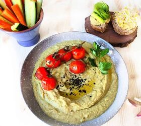 easy creamy all in one avocado chickpea hummus, A light blue bowl of avocado hummus topped with a drizzle of olive oil sprinkled with sesame seeds accompanied by six baked cherry tomatoes and a small parsley leaf Behind are two slices of bread one with avocado topping an the other with alfalfa sprouts There is oslo a small cup shape bowl with cucumber and carrot sticks