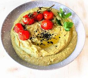 easy creamy all in one avocado chickpea hummus, A light blue bowl of avocado hummus topped with a drizzle of olive oil sprinkled with sesame seeds accompanied by six baked cherry tomatoes and a small parsley leaf