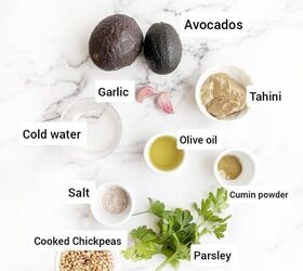 easy creamy all in one avocado chickpea hummus, An overhead view of various ingredients for making avocado hummus arranged on a marble surface The ingredients include two ripe avocados a few garlic cloves tahini olive oil cooked chickpeas cold water salt cumin powder and fresh parsley Each ingredient is labeled clearly for easy identification