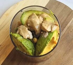 easy creamy all in one avocado chickpea hummus, A food processor bowl filled with whole ingredients for avocado hummus ripe avocado halves chickpeas garlic cloves tahini and a sprinkle of cumin ready to be blended into a creamy dip