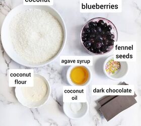 chocolate bounty blueberry bites, Ingredients for Chocolate Bounty Blueberry Bites laid out on a white marble surface including shredded coconut blueberries fennel seeds agave syrup coconut flour coconut oil and dark chocolate