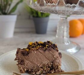 no bake chocolate orange mousse cake with crunchy oats, A slice of no bake chocolate orange mousse cake on a white plate a small gold fork lays in front The whole cake is displayed on a glass stand in the background with fresh oranges and flowers