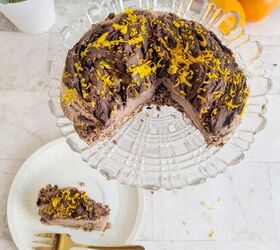 no bake chocolate orange mousse cake with crunchy oats, A top down view of a no bake chocolate orange mousse cake on a glass cake stand garnished with dark chocolate chunks and vibrant orange zest A slice is served on a white plate below with a golden fork beside it Fresh oranges and potted plants decorate the background