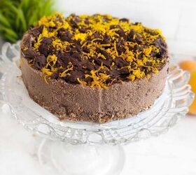 no bake chocolate orange mousse cake with crunchy oats, A close up of a no bake chocolate orange mousse cake on a glass cake stand featuring a smooth rich chocolate mousse layer a textured nutty base and a topping of dark chocolate chunks with bright orange rind The blurred background includes green plants and fresh oranges