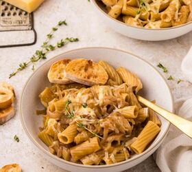 french onion pasta, A bowl of rigatoni pasta topped with grated cheese caramelized onions fresh thyme and toasted bread slices with another bowl and cheese grater in the background on a light surface