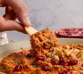 muhammara recipe roasted red pepper dip gf, A hand dips pita bread into a vibrant muhammara recipe the red pepper dip adorned with pomegranate seeds chopped walnuts and parsley A halved pomegranate sits in the background