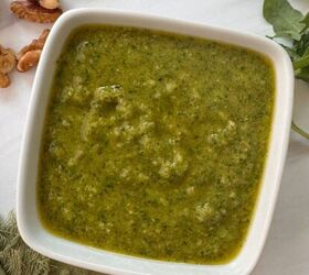 muhammara recipe roasted red pepper dip gf, A square white bowl filled with green walnut pesto sauce sits on a white surface with arugula leaves and walnut pieces nearby A green cloth and silver spoon are also partially visible next to the bowl