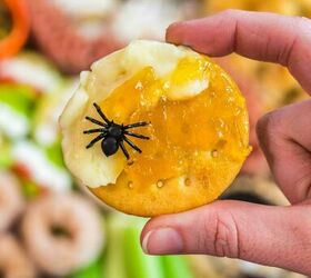 spooky sweet the ultimate halloween charcuterie board, A hand holds a round cracker topped with cheese orange jam and a small plastic black spider with a colorful snack platter blurred in the background