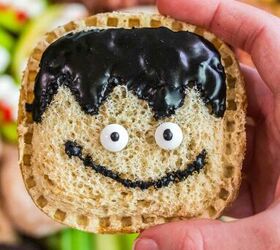 spooky sweet the ultimate halloween charcuterie board, A hand holds a square sandwich with a whimsical face made from candy eyes and black icing shaped like hair and a smile resembling a cartoon character The background features assorted snacks