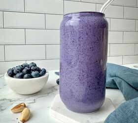blueberry protein shake, A purple smoothie in a clear glass A bowl of blueberries Brazil nuts and chia seeds are on a marble table with a blue kitchen towel
