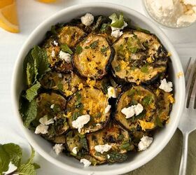 mediterranean roasted zucchini slices, A white bowl filled with roasted zucchini slices garnished with crumbled cheese fresh mint chopped herbs and lemon zest Orange wedges and a small dish of cheese rest nearby on a white surface
