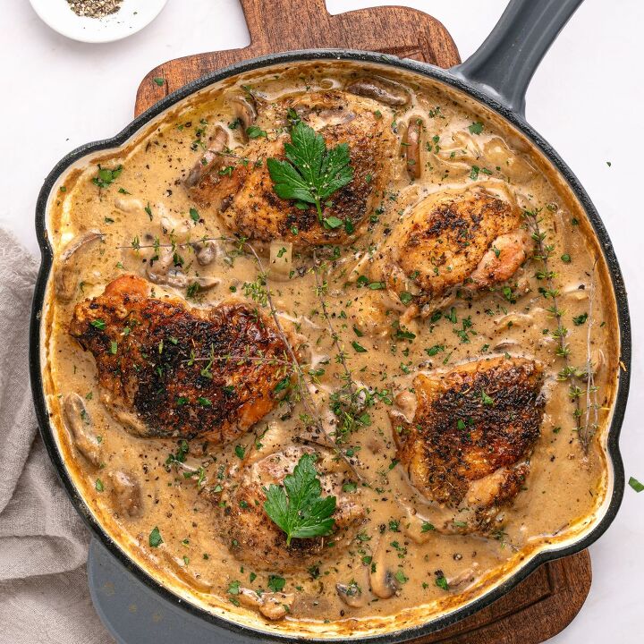 juicy chicken marsala thighs gf one skillet, A skillet filled with four browned chicken thighs in a creamy mushroom sauce inspired by Chicken Marsala garnished with fresh parsley and herbs sits on a wooden board with a small dish of ground pepper nearby