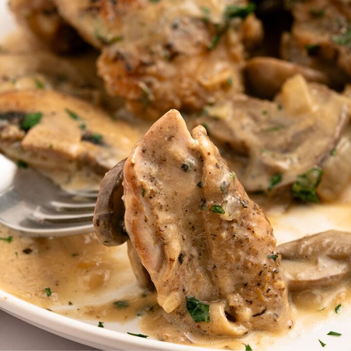 juicy chicken marsala thighs gf one skillet, A close up of a fork holding a piece of chicken marsala covered in creamy mushroom sauce garnished with chopped herbs with more saucy chicken thighs and mushrooms on a white plate all made in one skillet in the background