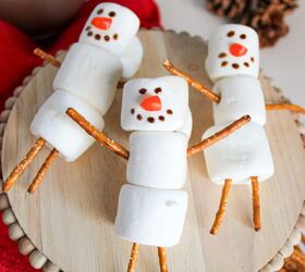 cute and easy marshmallow snowmen for the holidays, Three marshmallow snowmen with pretzel stick arms and legs arranged on a wooden stand surrounded by a red towel hot cocoa and pinecones