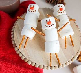 cute and easy marshmallow snowmen for the holidays, Close up of marshmallow snowmen with pretzel stick limbs and decorated faces on a wooden tray next to a glass of hot chocolate