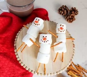 cute and easy marshmallow snowmen for the holidays, Far view of marshmallow snowmen with pretzel stick limbs and decorated faces on a wooden tray next to a mug of hot chocolate