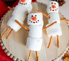 cute and easy marshmallow snowmen for the holidays, Top view of marshmallow snowmen with pretzel arms and legs festive red towel and hot chocolate in the background