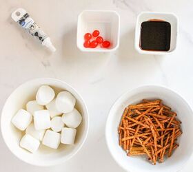 cute and easy marshmallow snowmen for the holidays, Marshmallows pretzel sticks red jelly beans chocolate syrup and black decorating gel arranged in bowls on a white marble surface